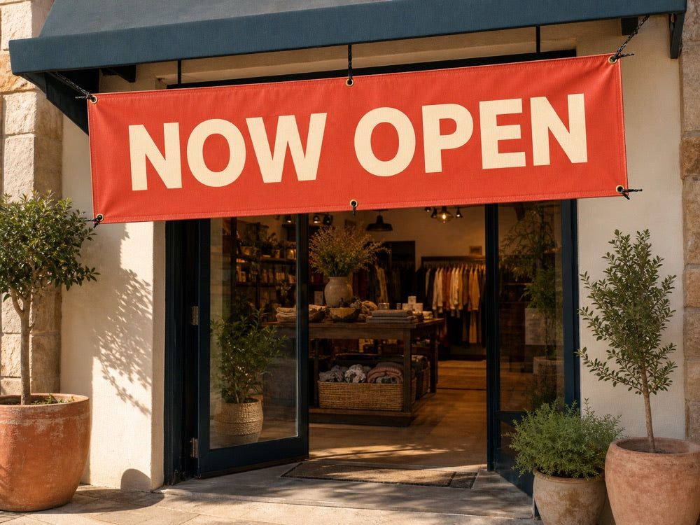 NOW OPEN vinyl banner hung from a boutique awning above an open shop door